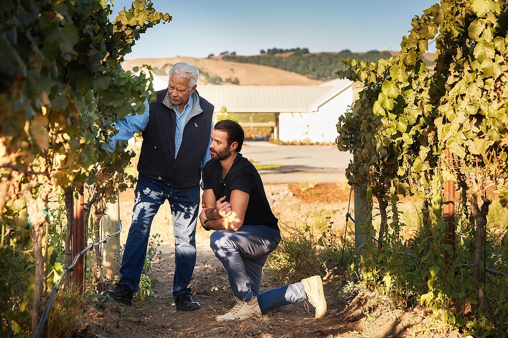 John Sweazey and John Michael standing between two vines in the J McK Estate Vineyard, blooming vines in the foreground, a building in the mid-ground, with hills of trees in the background. 
